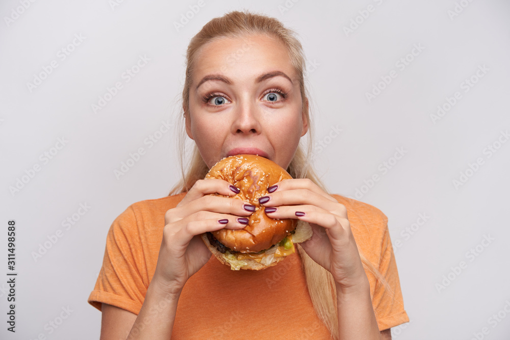 Close-up of overjoyed young blue-eyed cute blonde female eating tasty burger with great pleasure and looking happily at camera with wide eyes opened, isolated over white background