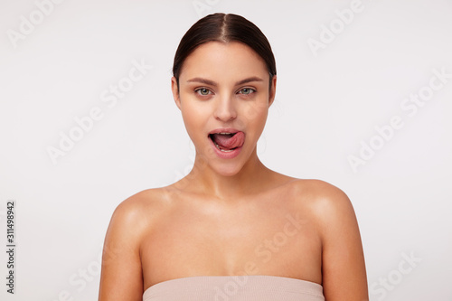 Frisky young attractive dark haired female with natural makeup looking playfully at camera and sticking out her tongue, dressed in beige top while posing over white background