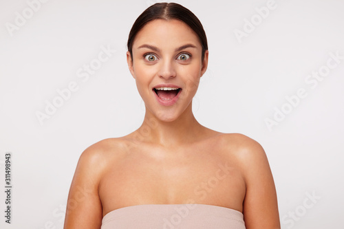 Portrait of surprised young cute brunette woman with casual hairstyle rounding eyes while looking at camera and keeping mouth wide opened, isolated over white background