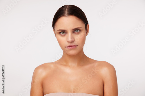 Close-up of gloomy young dark haired green-eyed lady with casual hairstyle looking seriously at camera and keeping lips folded, posing against white background with naked shoulders
