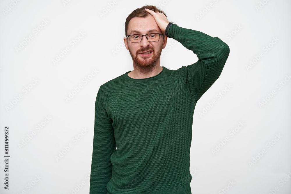 Fototapeta premium Bewildered young pretty brunette bearded male wearing glasses while standing over white background, clutching his head wih raised hand and twisting confusedly mouth