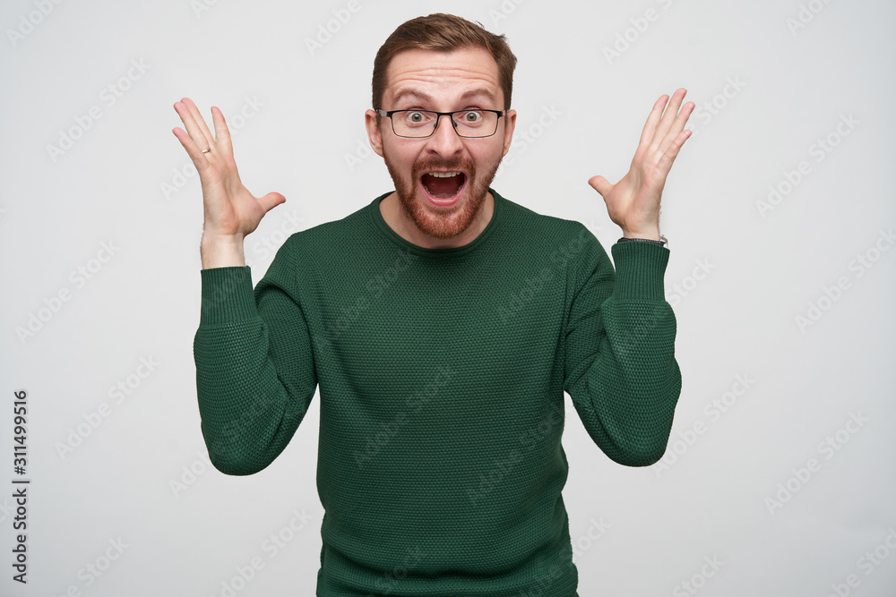 Naklejka premium Overjoyed young pretty brunette bearded male in glasses raising happily hands and looking at camera with wide eyes and mouth opened, standing against white background
