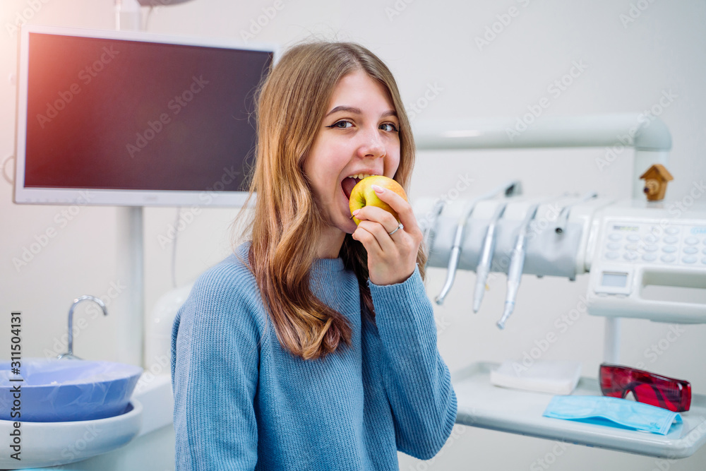 Happy girl sitting in dental chair and eating fresh apple after successful dental treatment. Closeup Happy girl sitting in dental chair and eating fresh apple after successful dental treatment. Closeup