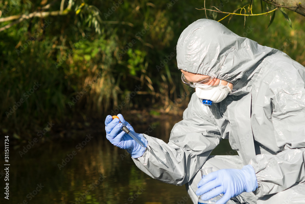 a man in a protective suit and mask holds a pipette of water from the river in his hands to pour into a test tube to study the quality of water