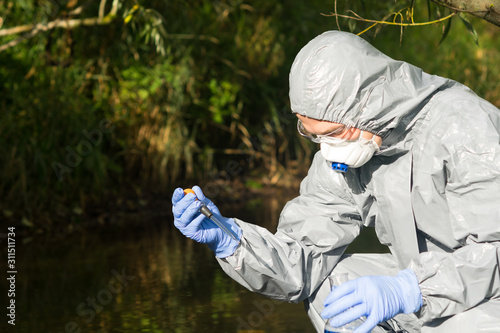 a man in a protective suit and mask holds a pipette of water from the river in his hands to pour into a test tube to study the quality of water