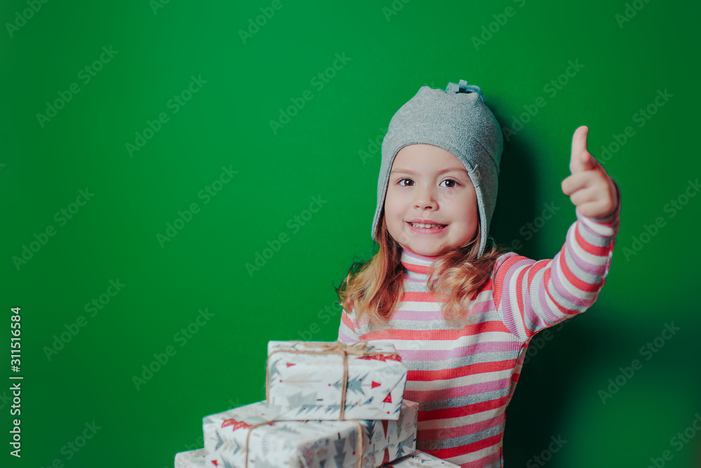 Christmas card with baby girl on the green background with copy space. A kid with christmas gifts. A child with many gift boxes shows thumb up. Christmas card with baby girl on the green background with copy space. A kid with christmas gifts. A child with many gift boxes shows thumb up.