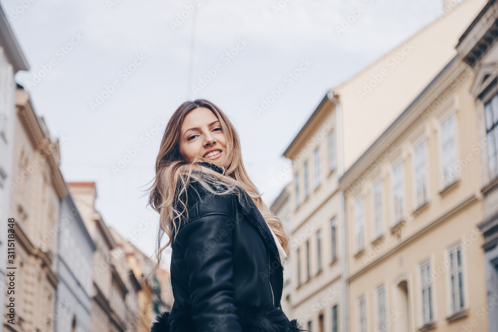 street style portrait of european attractive blonde woman wearing leather trousers and jacket. crossing the street and smiling at camera. fashion outfit details perfect for autumn fall winter