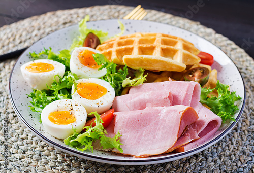 Breakfast with cornmeal waffles, boiled egg, ham and tomato on dark background. Appetizers, snack, brunch. Healthy food.