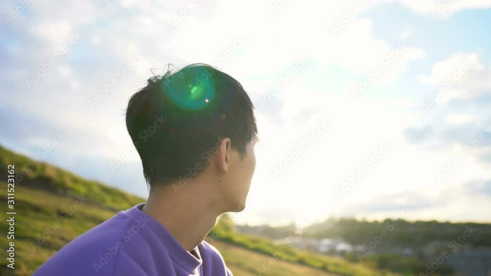 Portrait of Happy 20s Asian Man Wearing Purple Hoodie Sitting on Grass Outdoors in Nature During Summer Afternoon