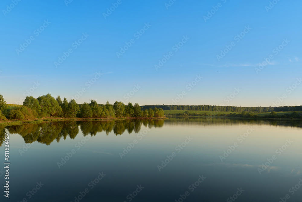 calm expanse of pond water on a bright sunny day with trees with green leaves on the opposite bank.