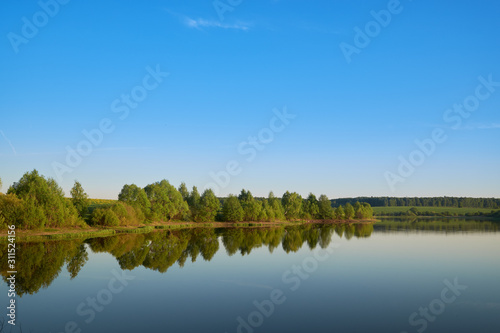 Fototapeta Naklejka Na Ścianę i Meble -  calm expanse of pond water on a bright sunny day with trees with green leaves on the opposite bank.