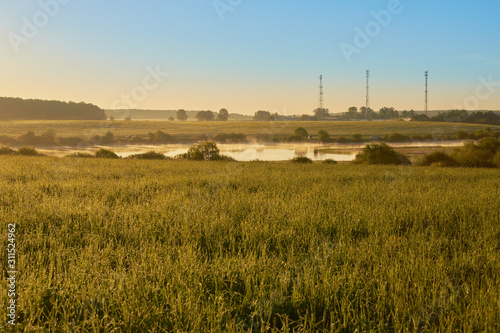 dawn with a view of the field and the river with three cell towers on the shore with clouds in the sky.
