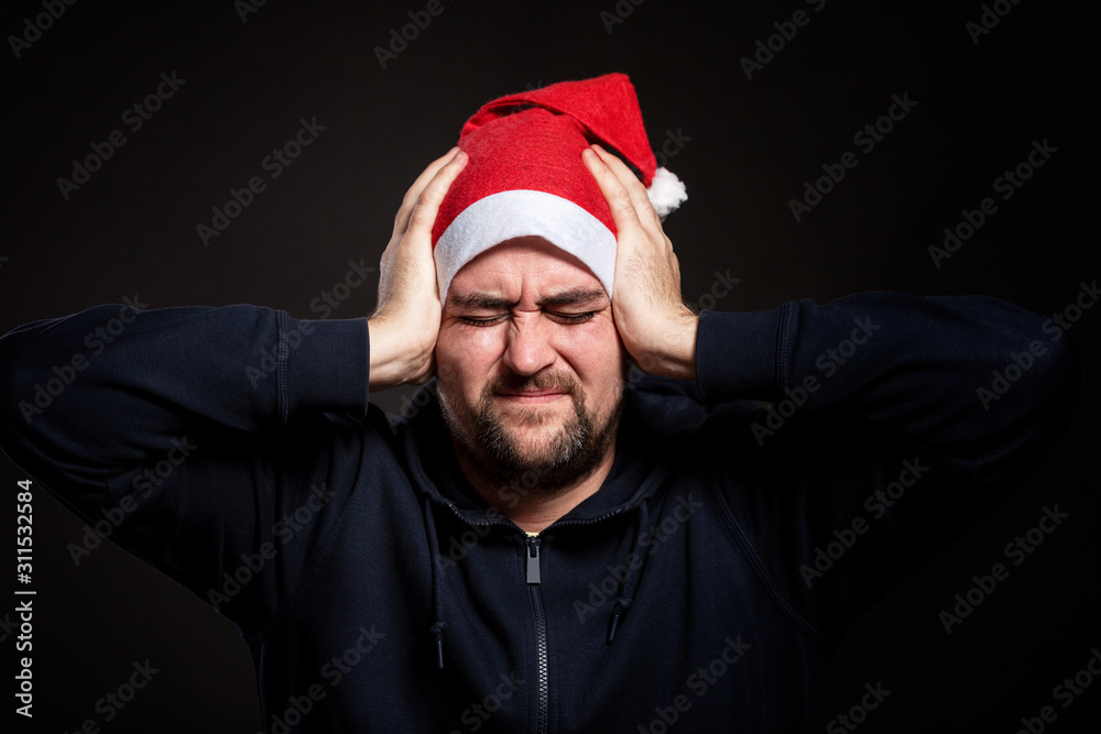 A handsome young man in a Santa Claus hat clasps his head with his hands and squints. Black background. Close-up. A handsome young man in a Santa Claus hat clasps his head with his hands and squints. Black background. Close-up.