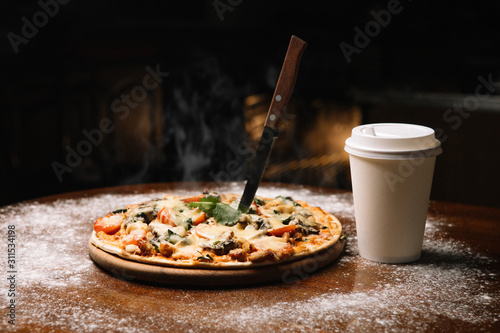 Hot large pizza and a glass of coffee tea pizza composition with melting cheese chicken tomatoes mushrooms steam smoke on a black background