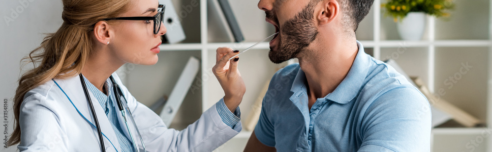 Obraz premium panoramic shot of attractive doctor in glasses examining man with medical spatula