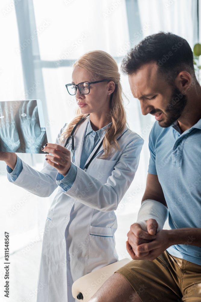 Obraz premium attractive orthopedist in glasses holding x-ray near injured man suffering from pain