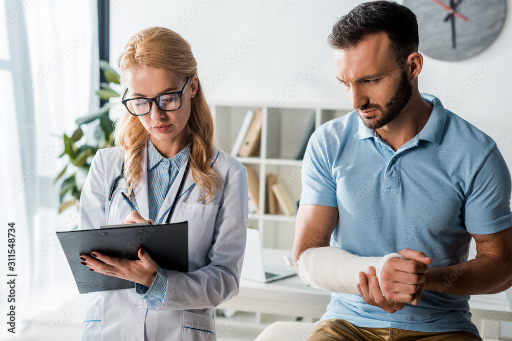 attractive orthopedist holding clipboard near injured bearded man attractive orthopedist holding clipboard near injured bearded man