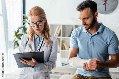 attractive orthopedist holding clipboard near injured bearded man