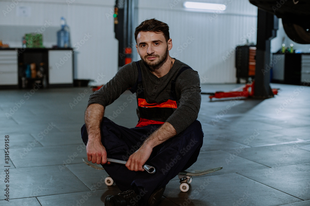 A young mechanic is sitting on a skateboard in a car service after finishing work smiling A young mechanic is sitting on a skateboard in a car service after finishing work smiling
