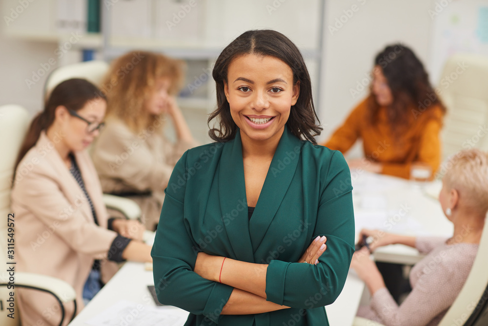 Charming African American woman standing with arms closed looking at camera smiling with colleagues coworking behind her, waist up portrait