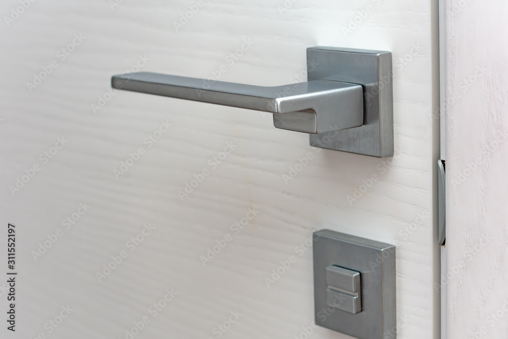 Modern chrome door handle and lock on white wooden door. Close-up elements of the modern interior of the apartment. Ajar white door. Modern chrome door handle and lock on white wooden door. Close-up elements of the modern interior of the apartment. Ajar white door.