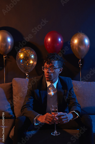 Portrait of trendy African-American man holding glass of champagne while sitting on sofa in dimly lit room at night club, copy space