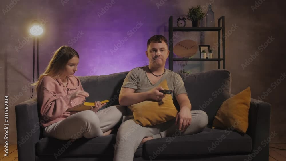 A man watches TV. Beautiful girl reading a book near to her husband A man watches TV. Beautiful girl reading a book near to her husband