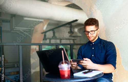 Handsome businessman distracted from work on the laptop watching video on smartphone. Freelancer holding mobile phone and browsing using high speed 4g or 5g internet. Man playing mobile games at work