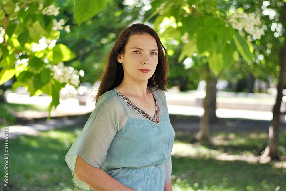 Beautiful brunette woman with long hair and blue eyes walking in the blooming park at spring in sunny day with bright sunshines and green trees Beautiful brunette woman with long hair and blue eyes walking in the blooming park at spring in sunny day with bright sunshines and green trees