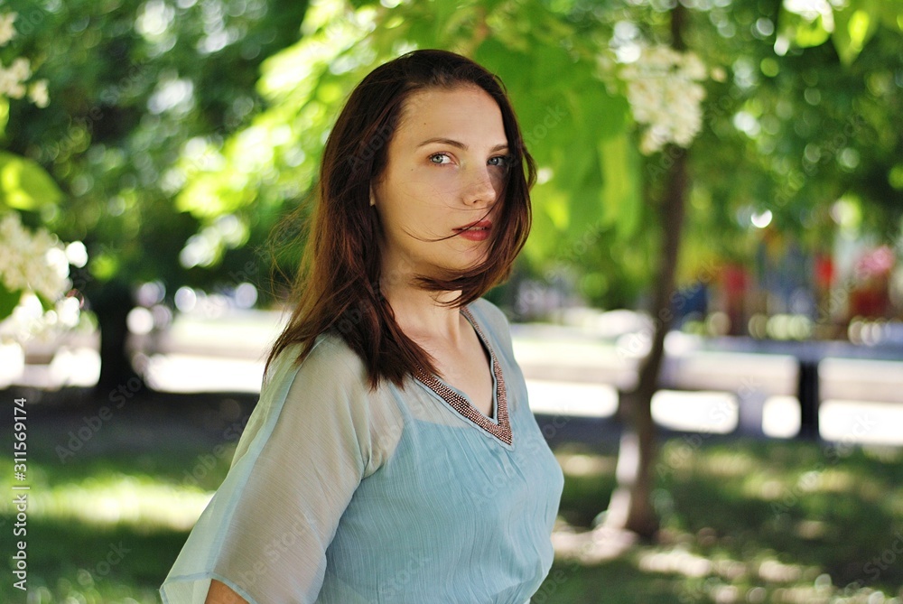 Beautiful brunette woman with long hair and blue eyes walking in the blooming park at spring in sunny day with bright sunshines and green trees Beautiful brunette woman with long hair and blue eyes walking in the blooming park at spring in sunny day with bright sunshines and green trees