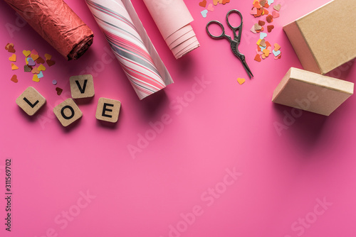top view of valentines decoration, scissors, gift box, wrapping paper and love lettering on wooden cubes on pink background