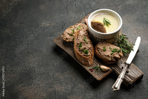 Fresh homemade chicken liver pate in ceramic bowl or ramekin and baguette slices with herbs and garlic on a dark background.