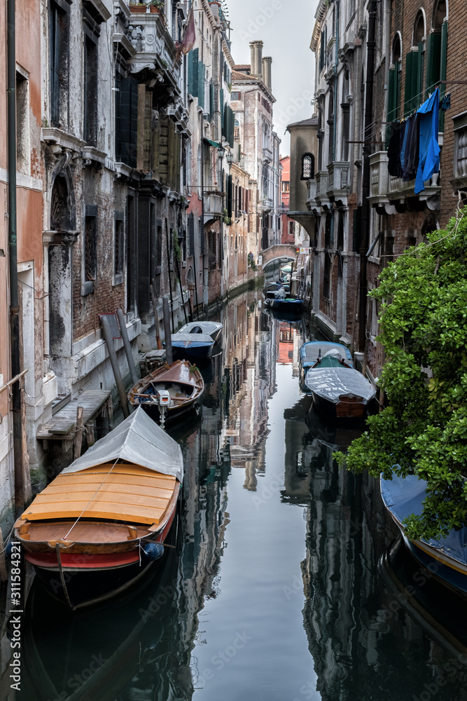Narrow canal between old houses, boats on dark water. Arch bridge. Venice, Italy.