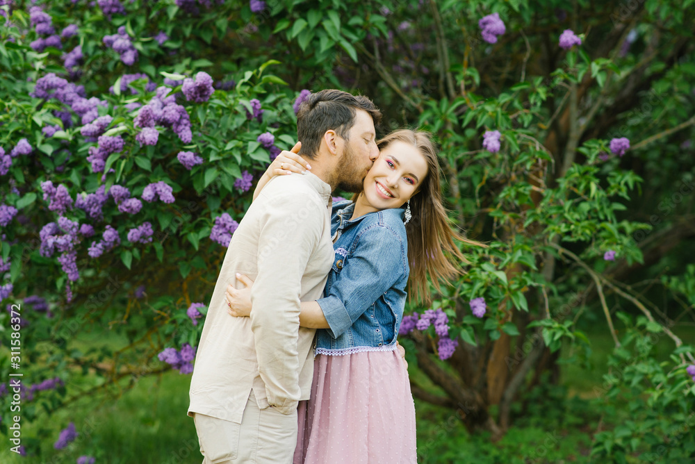 Happy couple in love in blooming lilac gardens in spring. The guy kisses the girl on the cheek, who smiles. Valentine's day Happy couple in love in blooming lilac gardens in spring. The guy kisses the girl on the cheek, who smiles. Valentine's day