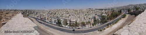 Cityscape of Amman, capitol of Jordan, grey panorama of a modern Arabic city with improvised houses on a hill between few green trees and a Roman theatre under the blue sky 