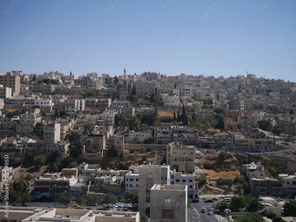 Cityscape of Amman, capitol of Jordan, grey panorama of a modern Arabic city with improvised houses on a hill between few green trees and a Roman theatre under the blue sky	