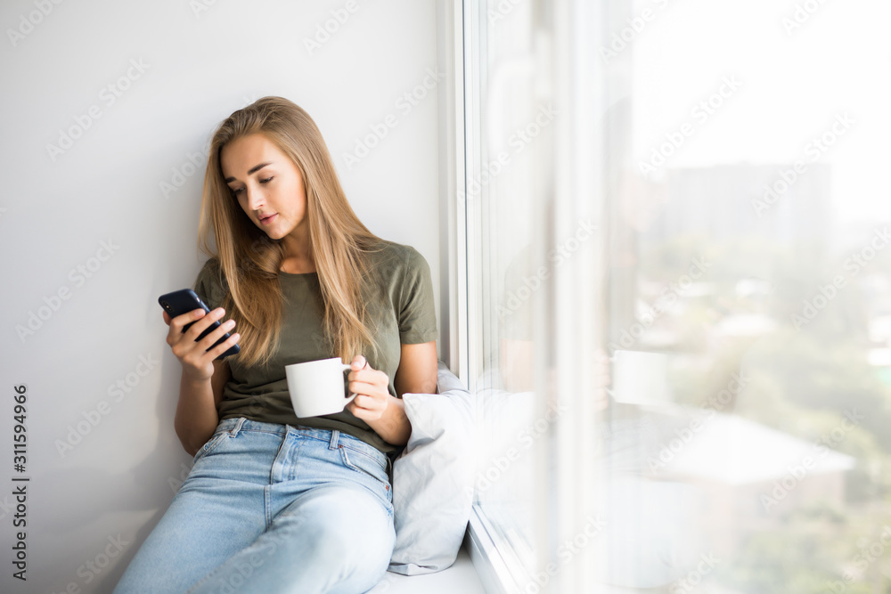 Beautiful young woman drinking coffee use mobile phone and looking through window while sitting at windowsill at home Beautiful young woman drinking coffee use mobile phone and looking through window while sitting at windowsill at home