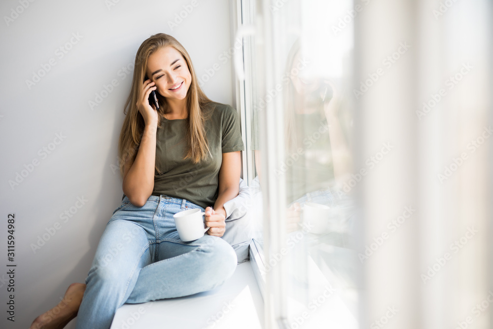 Young smiling woman talking on the mobile phone while sitting on the windowsill Young smiling woman talking on the mobile phone while sitting on the windowsill