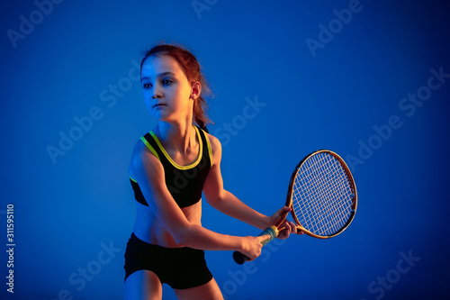 Little caucasian girl playing tennis on blue studio background in neon light. Cute model training in motion, action. Youth, flexibility, power, energy. Movement, ad, sport, healthy lifestyle concept.