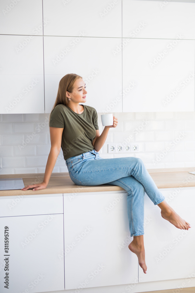 Young pretty woman holding cup with coffee or tea while sitting on counter in the kitchen