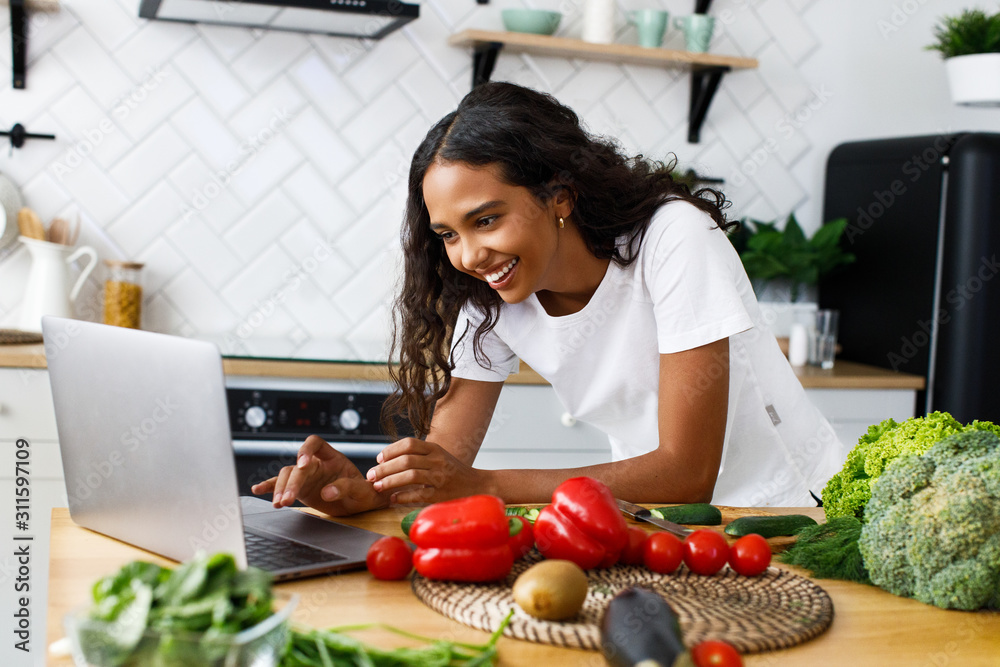 Smiled pretty girl is looking on the laptop screen on the modern kitchen on the table full of vegetables and fruits, dressed in white t-shirt Smiled pretty girl is looking on the laptop screen on the modern kitchen on the table full of vegetables and fruits, dressed in white t-shirt