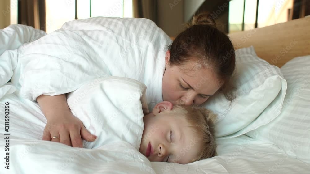 A caring mother kisses her sleeping son in the morning in bed, she wakes him up. A high resolution. Mom and son are sleeping in a white bed in the morning, they woke up.