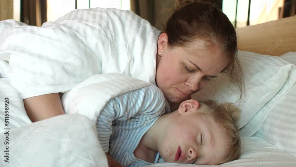 A caring mother kisses her sleeping son in the morning in bed, she wakes him up. A high resolution. Mom and son are sleeping in a white bed in the morning, they woke up. A caring mother kisses her sleeping son in the morning in bed, she wakes him up. A high resolution. Mom and son are sleeping in a white bed in the morning, they woke up.