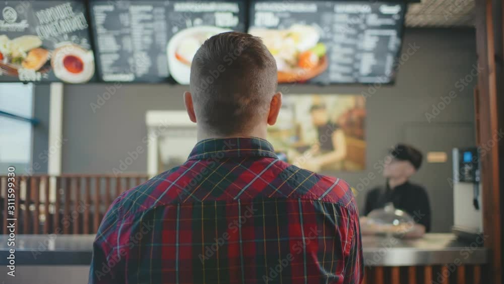 Back shot male person near cash desk in fastfood cafe. Man in casual wear checkered shirt inside food restaurant looking to menu board choosing dish to eat