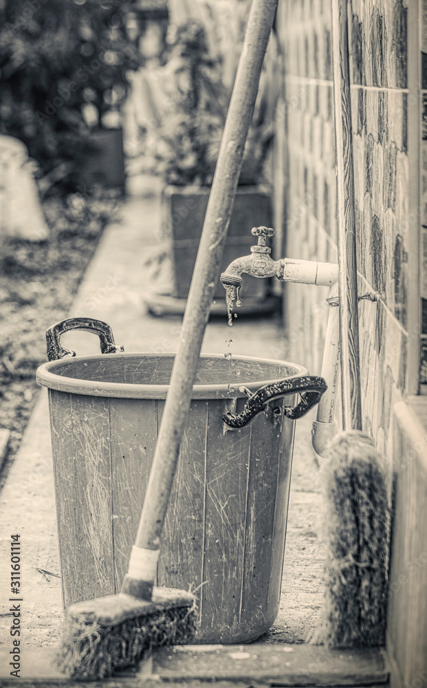 Old and dirty cleaning equipments in a garden. Plastic bucket and brooms. Old and used Vintage open faucet. Black and white photography Old and dirty cleaning equipments in a garden. Plastic bucket and brooms. Old and used Vintage open faucet. Black and white photography