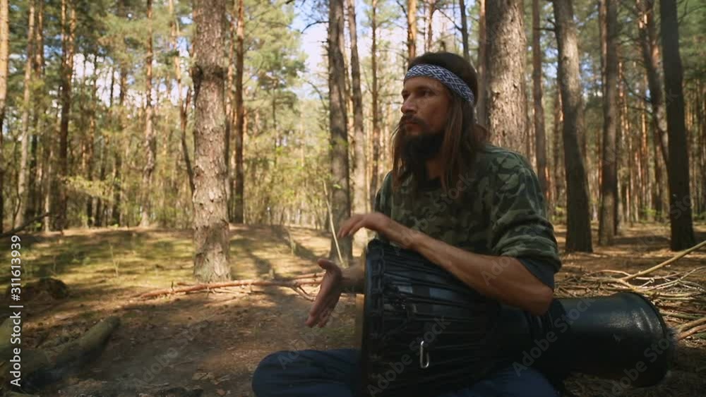 Young Caucasian hippie man playing a djembe drum in the summer coniferous forest Young Caucasian hippie man playing a djembe drum in the summer coniferous forest