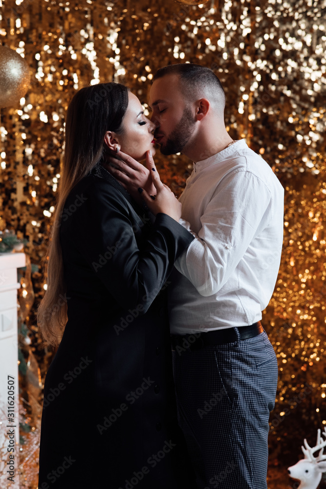 Woman in black dress with nice hair hug and kiss her husband on christmas eve. Hot of a young happy couple in love celebrating Christmas. Holidays and celebration concept.