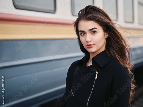 Young beautiful brown haired woman in black jacket blue skirt with gorgeous long legs in vintage beret emotionally posing on railway station train background with cinematic color grading photo