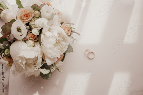 Wedding bouquet with beautiful roses and pions with 2 wedding rings lying on the floor near window with curtains. Long shadow of the window