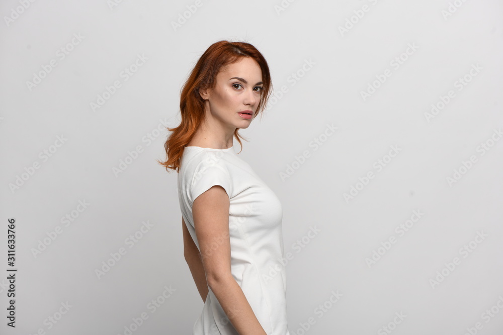 Beautiful redhead girl in a beige dress posing in front of the camera. Portrait on a neutral light background, space for text. Side view Beautiful redhead girl in a beige dress posing in front of the camera. Portrait on a neutral light background, space for text. Side view